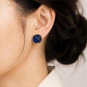 May include: A square stud earring with rounded corners featuring a deep blue textured surface with subtle gold-toned vein-like patterns. The earring is encased in a smooth gold-toned metal frame, creating a distinct contrast between the dark blue centre and the metallic border.