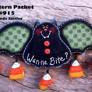 May include: A black bat with green checkered wings and red buttons, hanging from candy corn. The bat says "Wanna Bite?" and is a Halloween-themed craft project.