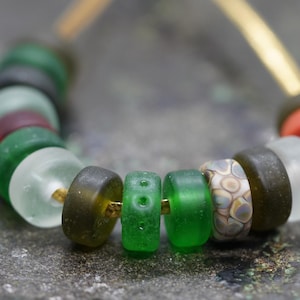 Pode incluir: Um colar de contas de vidro coloridas em tons de verde, castanho, branco e vermelho. As contas são enfiadas com um fio dourado. As contas são cilíndricas e têm uma textura lisa.