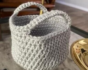 May include: A small, gray crocheted basket with a single handle. The basket is made with a textured stitch and has a rounded shape.