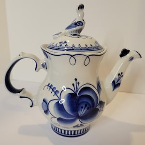 May include: White ceramic teapot with blue floral and scroll designs. The teapot features a bird-shaped handle on the lid and a curved handle on the side. The spout is also decorated with blue accents. A decorative piece for the kitchen.