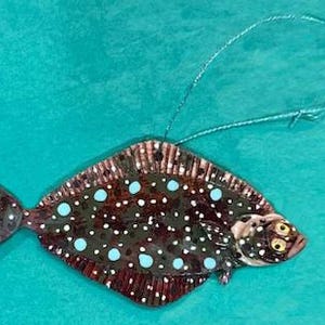 May include: A ceramic flounder ornament, hand-painted with a dark brown body, white and light blue spots, and a silver hanging string. The fish has yellow eyes and detailed fins. The ornament is a decorative item.