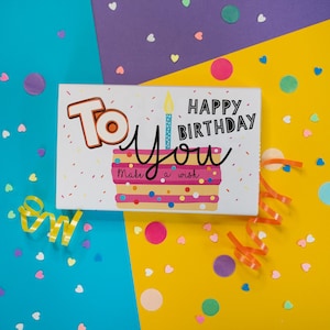 Pode incluir: Um cartão de aniversário com uma ilustração de bolo e as palavras "To You Happy Birthday". O cartão tem um fundo branco com um bolo colorido, confetes e uma vela acesa. O fundo é azul, roxo e amarelo.