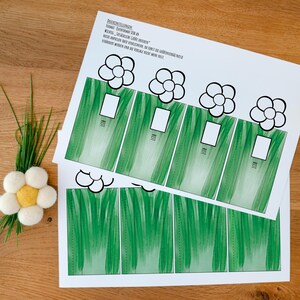 May include: A white paper sheet featuring a printed template of green grass blades and black-outlined flower shapes, with text in German at the top. Beside the paper is a small, handmade white and yellow felt flower with green stems.