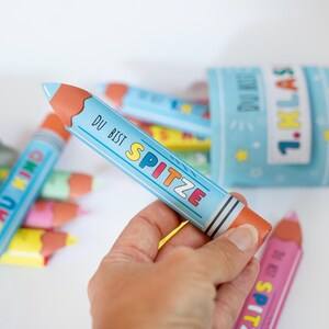 May include: A light blue pencil-shaped container with the words "DU BIST SPITZE" in colorful letters. The pencil has a brown tip and is held in a hand. Other similar pencils and a container with "1. KLAS" are in the background.