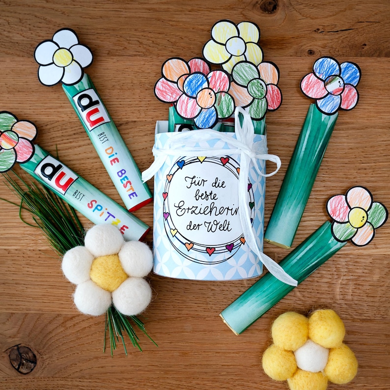 May include: A cylindrical container with a light blue geometric pattern and a white ribbon, featuring the text "F&uuml;r die beste Erzieherin der Welt". It is surrounded by green tubes topped with hand-colored paper flowers and two felt flowers.