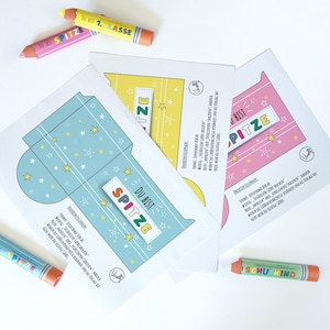 May include: Three colorful paper cards with star and stripe designs, each featuring the text "DU BIST SPITZE". Several crayon-shaped pencils are scattered around the cards. The pencils have text on them.