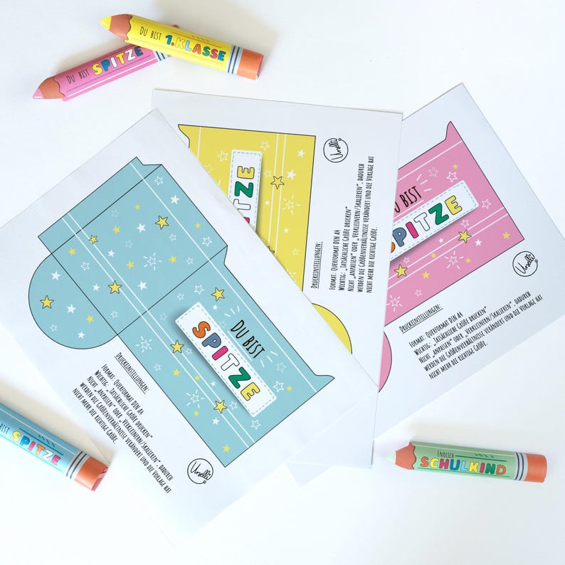 May include: Three colorful paper cards with star and stripe designs, each featuring the text "DU BIST SPITZE". Several crayon-shaped pencils are scattered around the cards. The pencils have text on them.