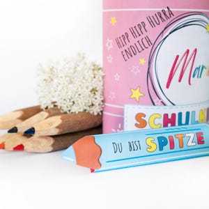 May include: A pink cylindrical container with the text "HIPP HIPP HURRA ENDLICH" and the name "Mari" in pink script. A blue pencil-shaped card reads "DU BIST SPITZE". Several pencils and white flowers are also present.