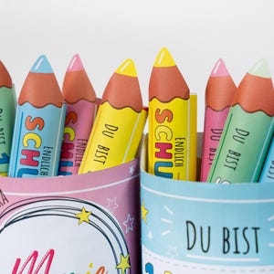 May include: A close-up of colorful pencil-shaped containers filled with decorative pencils. The pencils are yellow, blue, pink, and green with the words "DU BIST" and "ENDLICH SCHU" printed on them. The containers are decorated with stars and the word "Marie".