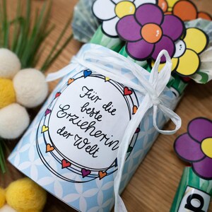 May include: A light blue cylindrical container with a white geometric pattern, tied with a white ribbon. A circular label features the German text "F&uuml;r die beste Erzieherin der Welt" surrounded by small colorful hearts. Paper flowers in purple, yellow, and white are attached.