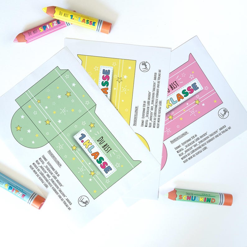 May include: Three colorful paper cutouts with the text "DU BIST 1.KLASSE" and star decorations, along with three pencil-shaped crayons. The cutouts are in green, yellow, and pink. The crayons are orange, pink, and blue.