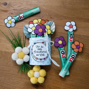 May include: A cylindrical container with a white ribbon and the text "F&uuml;r die beste Erzieherin der Welt" surrounded by paper flowers made from currency, accompanied by green tubes labeled "du bist gro&szlig;artig" topped with paper flowers and felt flowers.