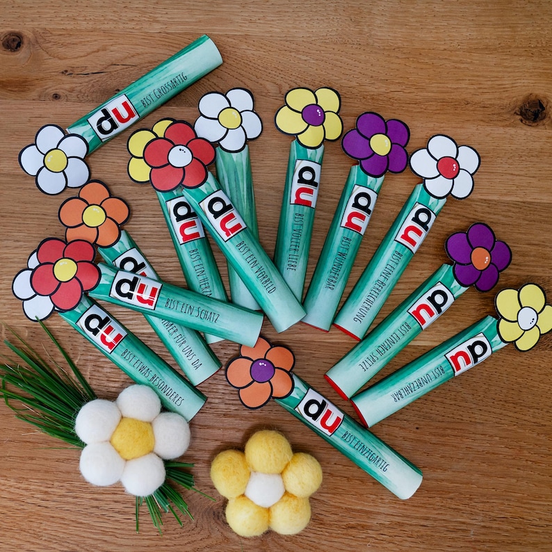 May include: A collection of light green cylindrical tubes, each topped with a colorful paper flower. The tubes feature the text 'du' followed by German phrases like 'Bist grossartig' and 'Bist ein Schatz'. Two felt flowers are placed below the tubes.