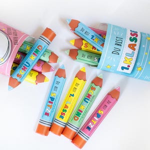 May include: Colorful pencil-shaped party favors with text in German. The pencils are blue, yellow, green, and pink, with orange erasers and brown tips. The pencils are scattered around two blue and pink paper cones.