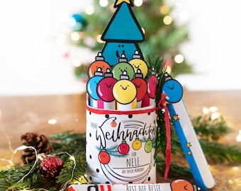 May include: A Christmas gift set featuring a cylindrical container with a Christmas tree and bauble design, a bookmark, and a candy bar. The container has the text "Frohe Weihnachten" and "Für Nele".