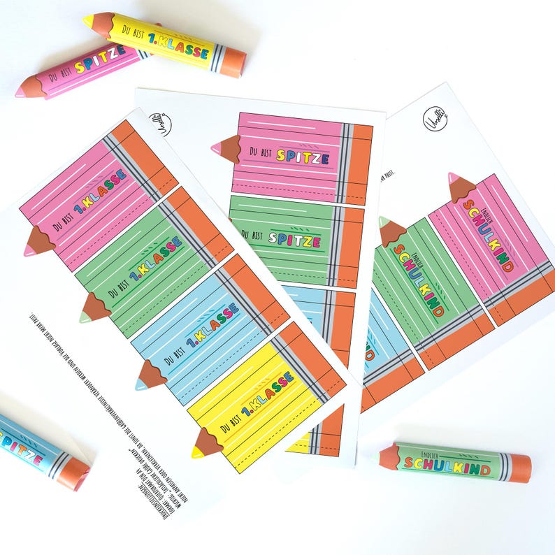 May include: A collection of colorful pencil-shaped cards and crayons. The cards feature a pencil design with the text "DU BIST 1.KLASSE" and "SPITZE". The crayons are cylindrical with the text "ENDLICH SCHULKIND". The color palette includes pink, green, blue, and yellow.