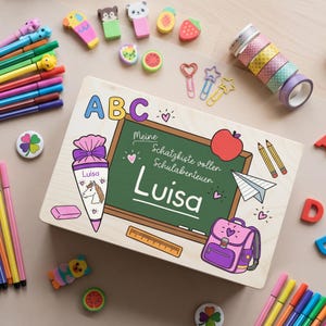Könnte beinhalten: Eine rechteckige Holzbox mit einem aufgedruckten grünen Tafel-Design mit dem Text "Meine Schatzkiste voller Schulabenteuer Luisa", einer rosa Schultüte, einem rosa Schulranzen, einem Apfel, einem Papierflieger und Stiften, umgeben von bunten Stiften.