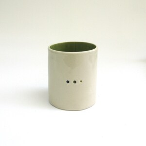 May include: A white ceramic cup with a green rim and three small holes. The holes are arranged in a row and are black, gray, and green.