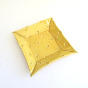 May include: A yellow ceramic square dish with a raised polka dot pattern. The dish has a slightly curved edge.