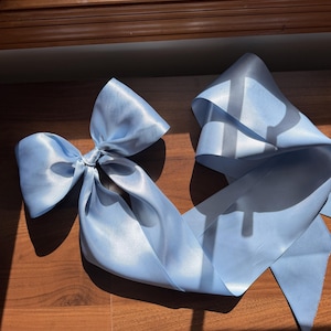 May include: A large, light blue satin hair bow featuring a smooth, reflective finish and long, flowing ribbon tails. The fabric has a soft sheen, and the bow is tied in a classic style with two prominent loops and two trailing ends.