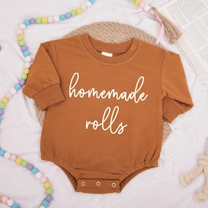 Pode incluir: Um macacão de bebê de manga comprida marrom com o texto "homemade rolls" em escrita branca.
