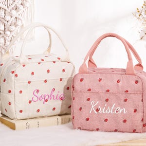Könnte beinhalten: Zwei Lunch-Taschen aus Cord, eine cremefarbene und eine rosa, sind mit roten Erdbeermustern verziert. Die cremefarbene Tasche hat den Namen "Sophia" in Rosa gestickt, die rosa Tasche "Kristen" in Weiß.
