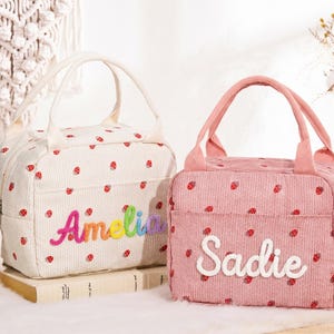 May include: Two rectangular lunch bags with top handles, one in cream and one in pink, both featuring a small red strawberry pattern. The cream bag has the name Amelia embroidered in rainbow colors, and the pink bag has the name Sadie in white.