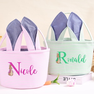 Puede incluir: Dos cestas de Pascua con orejas de conejo. Una es rosa con rayas blancas y el nombre "Nicole" en rosa. La otra es verde con rayas blancas y el nombre "Ronald" en verde. Ambas tienen un gráfico de Peter Rabbit.