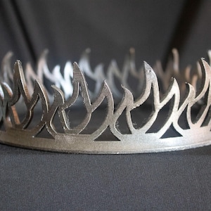 May include: A silver crown with a flame design. The crown is made of metal and has a smooth finish.