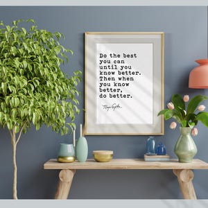 May include: A framed print with the quote "Do the best you can until you know better. Then when you know better, do better." is displayed above a wooden console table. The table holds decorative vases, bowls, and a small plant arrangement. A large green plant is on the left.