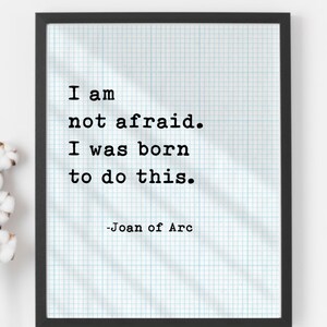 May include: A framed print with a black border features the quote "I am not afraid. I was born to do this." attributed to Joan of Arc. The text is in a bold, typewriter-style font on a grid-lined background.
