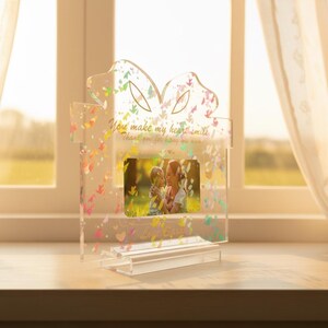 May include: Clear acrylic photo frame in a gift box shape. The frame has a butterfly design with colorful accents and the text "You make my heart smile". The frame holds a photo of a mother and child. The frame is on a clear base.