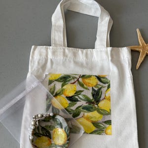 May include: A white tote bag with a lemon print, a small painted oyster shell dish, and a starfish. The tote bag has a rectangular lemon print on the front. The oyster shell dish is painted with lemons and pearls.