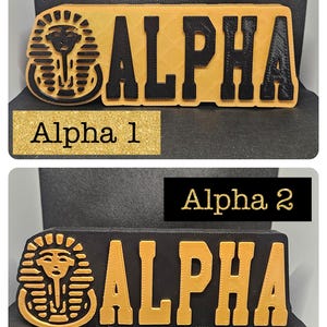 May include: Two versions of a black and gold wooden sign with the word "ALPHA" in gold letters. The sign also features a gold Egyptian pharaoh head with a cobra on the left side.