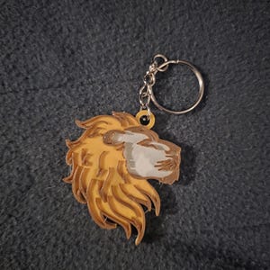 Könnte beinhalten: Ein Schlüsselanhänger mit einem Löwenkopf-Design. Die Löwenmähne ist in einem warmen Gelb-Orange gehalten, mit einem helleren, hellbraunen Gesicht und einem silbernen Schlüsselring. Der Schlüsselanhänger besteht aus 3D-gedrucktem Material.