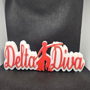 May include: White and red 3D sign with the words "Delta Diva" and a red silhouette of a woman dancing.