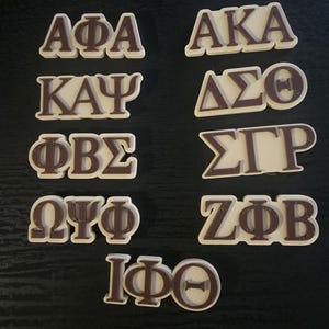 May include: A selection of brown and white Greek letters, including Alpha Phi Alpha, Alpha Kappa Alpha, Kappa Alpha Psi, Delta Sigma Theta, Phi Beta Sigma, Sigma Gamma Rho, Omega Psi Phi, Zeta Phi Beta, and Iota Phi Theta. The letters are displayed on a dark surface.