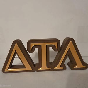 May include: A brown wooden Delta Tau Lambda sign with gold-colored letters. The letters are in a serif font and are raised from the base. The sign is likely used for decoration or display.