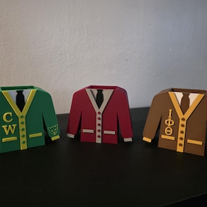 May include: Three decorative sweater-shaped containers in green, red, and brown. The green sweater has "CW" and a bear logo, the red has buttons, and the brown has Greek letters and "1963". Each has a white collar and black tie.