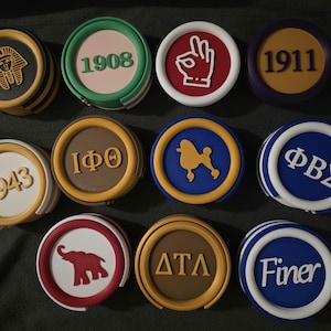 May include: A collection of round coasters with various designs and colors. Some coasters feature text like "1908", "1911", "1943", and "Finer". Other coasters have graphics including a hand gesture, an elephant, a poodle, and a pharaoh's head.