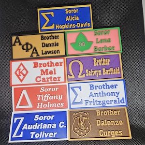 Può includere: Dieci targhette rettangolari colorate con bordi bianchi. Ogni targhetta presenta una lettera greca, un nome e un titolo, come "Soror" o "Brother".