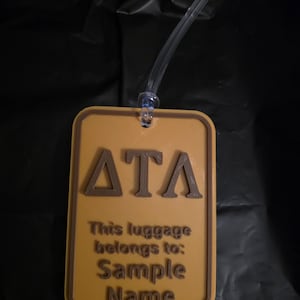 May include: A rectangular luggage tag with a brown border and text. The tag is tan with brown lettering that reads "ATA" at the top, and "This luggage belongs to: Sample Name" below. A clear plastic loop is attached at the top.
