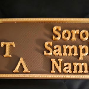 May include: A rectangular plaque with a brown background and gold-coloured trim. The plaque features the Greek letters Delta, Tau, and Lambda, along with the words "Soror Sample Name" in gold lettering. The background has a textured pattern.