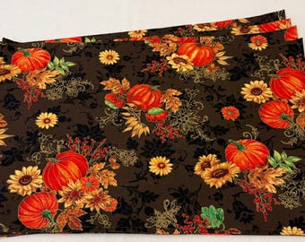 May include: A set of rectangular placemats featuring a dark brown background with a repeating pattern of orange pumpkins, yellow sunflowers, red berries, and autumn leaves. The fabric has a detailed print with gold-toned accents throughout.