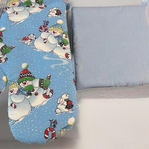 May include: A blue oven mitt and pot holder set. The oven mitt features a whimsical pattern of snowmen, dogs, and candy canes on a light blue background. The pot holder is a solid light blue color.