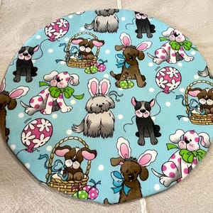 May include: A round, light blue fabric with a repeating pattern of cartoon dogs wearing bunny ears, polka dots, Easter eggs, and baskets. The dogs are in various colors, including brown, black, and white.