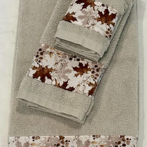 May include: A set of three beige terry cloth towels featuring a decorative fabric band with a printed pattern of brown and tan autumn maple leaves and small berries. The towels are stacked, displaying the textured fabric and the leaf design.
