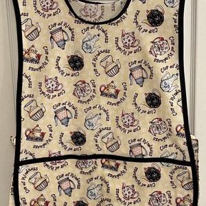 May include: A beige apron with a black trim and a large pocket. The apron is covered in a pattern of teapots and the words "Cup of Kindness".