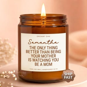 May include: A lit candle in a brown glass jar with a white label. The label reads "Samantha" and a message about motherhood. The candle is on a white surface with a soft pink background.
